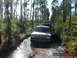 1989 Ford Bronco - Two trucks navigate a water-filled path in a wooded area.