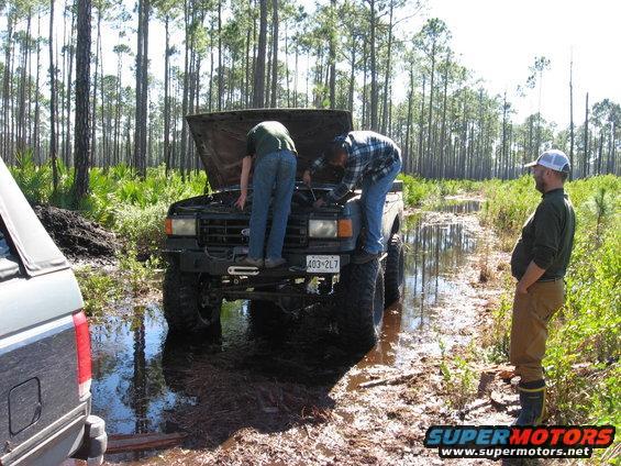 1989 Ford Bronco with hood open, stuck in mud, two people working on engine, another person watching