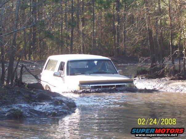 White 1989 Ford Bronco driving through muddy water, front 3/4 view