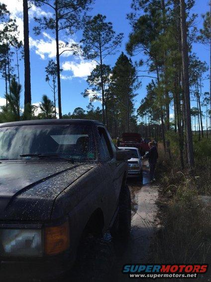 Front of a dark SUV, muddy, with other vehicles lined up on a dirt road in a forest