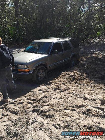 Silver SUV stuck in sand with a person standing nearby. The vehicle appears to be a Chevrolet Blazer from the late 1990s