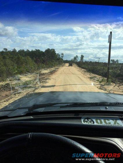 Dashboard view from inside a vehicle looking out at a dirt road with a distant SUV and trees under a cloudy sky
