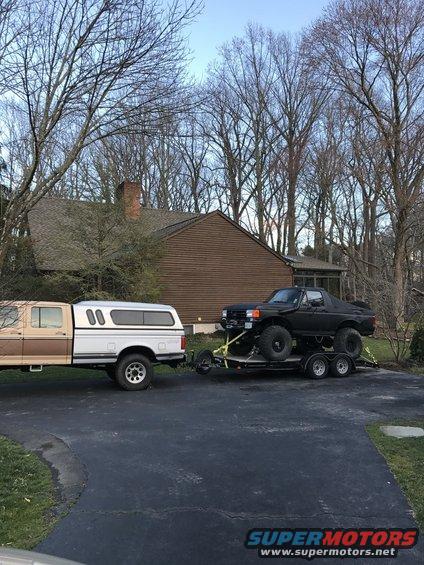 Brown and white pickup truck towing a black lifted truck on a trailer. The black truck appears to be a Ford Bronco