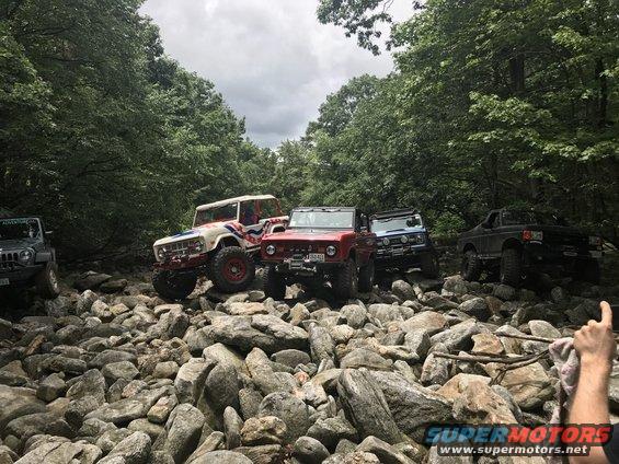 Several off-road vehicles, including a red Bronco and a white Bronco with red and blue stripes, navigate a rocky trail