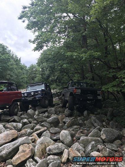 Three modified off-road trucks navigate a boulder-strewn path in a wooded area; one is red, one black, and one gray