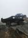 1989 Ford Bronco - Two off-road vehicles, a black Ford Bronco and a blue Jeep Wrangler, navigate a rocky incline on a foggy day.