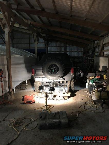 Gray Ford Bronco rear, lifted, with spare tire mounted. Underneath is being worked on in a dimly lit garage