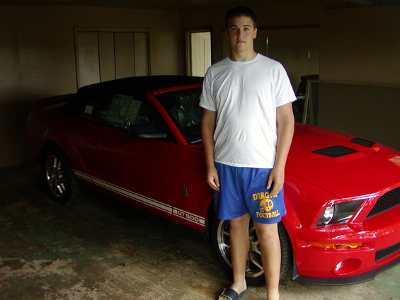 guy in white t-shirt and blue shorts stands next to a red 2007 Ford Mustang convertible with a black top