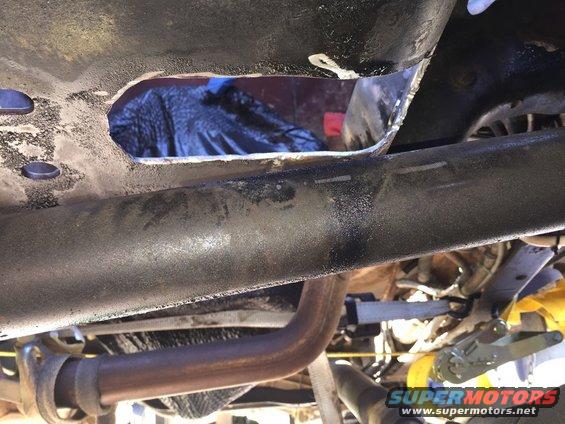 Close-up of a vehicle's underbody showing a large hole cut into the frame rail. Exhaust pipe visible below