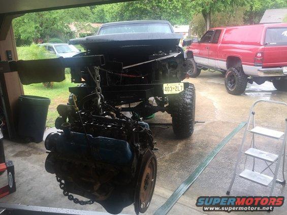 Engine hanging from a chain, with a truck chassis and a red pickup truck in the background