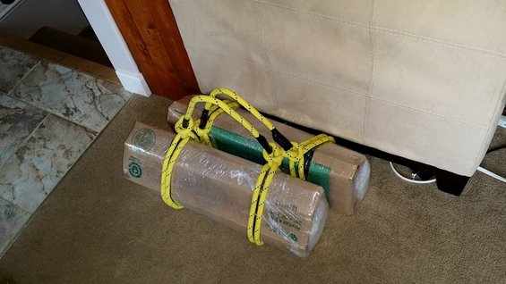 Two cardboard boxes wrapped in plastic and secured with yellow rope, resting on a carpet next to a couch
