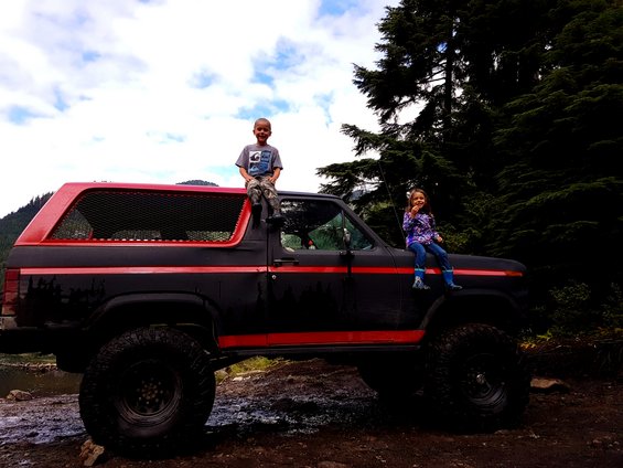 Two kids sit on a lifted black Ford Bronco with red trim. One child is on the roof, the other on the hood