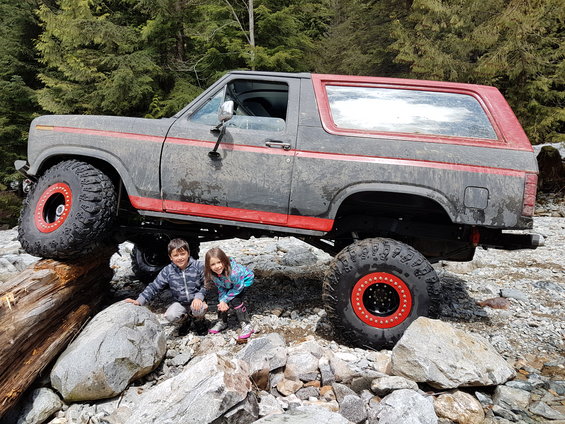 Lifted 1980 Ford Bronco with red trim and custom wheels, parked on rocks with two children