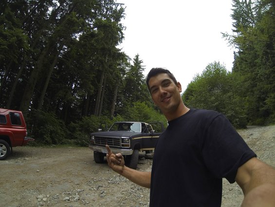Man in black t-shirt points at a dark-colored pickup truck parked on a dirt road, with a red SUV to the left. Trees surround the area