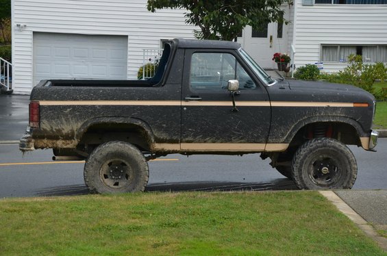Black Ford Bronco with tan stripe, covered in mud, parked on a wet street