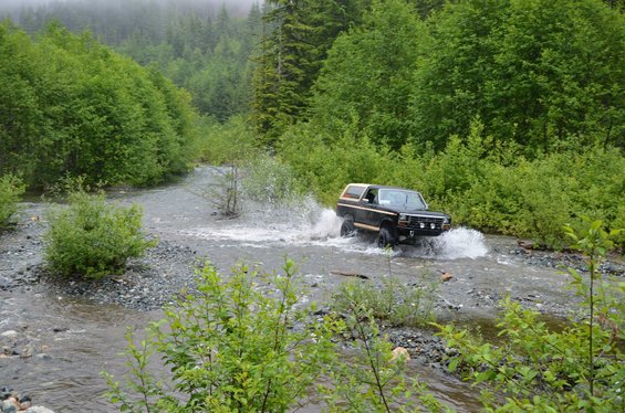 Black Ford Bronco driving through a shallow stream with trees and fog in the background