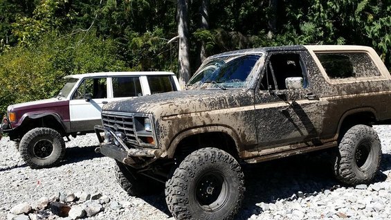 Muddy Ford Bronco and another SUV parked on a gravel road with trees in the background