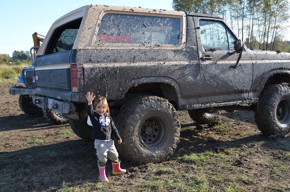Mud-covered 1980s Ford Bronco with a child waving in front. Another vehicle is partially visible behind it