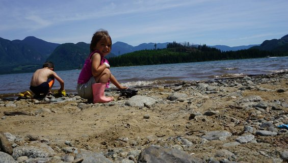 Two kids play on a beach by a lake with mountains in the background