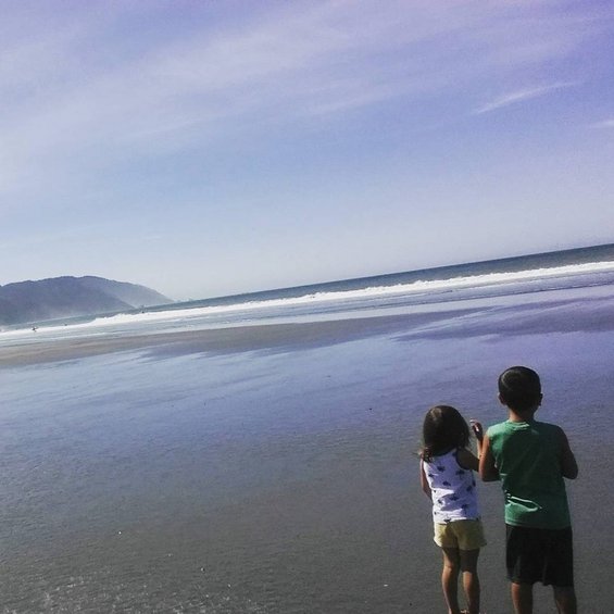 Two children stand on a wet sandy beach looking out at the ocean waves under a clear blue sky