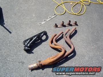 Exhaust manifolds, one black, one rusty, with gasket and bolts on asphalt