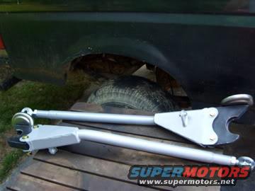 Two silver suspension arms on a wooden surface with a dark vehicle fender and tire in the background