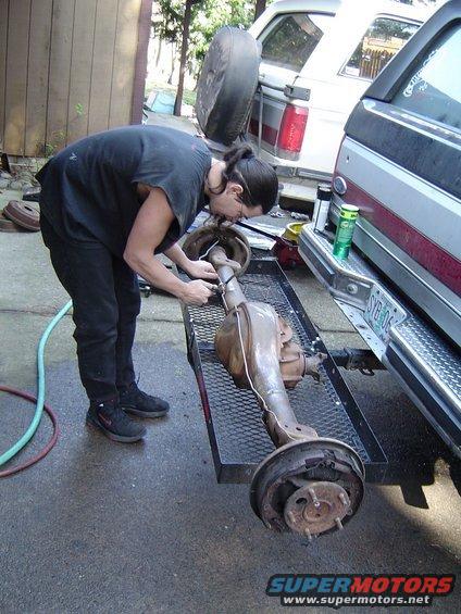 Person working on a rear axle assembly on a trailer, with a white SUV and a pickup truck in the background