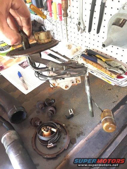 Fuel pump assembly with hoses, bolts, and a fuel sender unit on a workbench with tools in the background