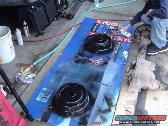 Two black brake drums on blue cardboard with spray paint cans and a person spraying. A differential is to the right