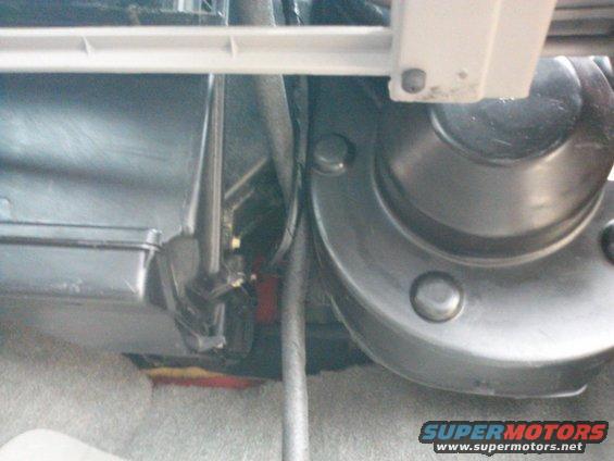 Close-up of vehicle interior parts, possibly near the dashboard or firewall. Shows a black plastic housing, metal components, and wiring