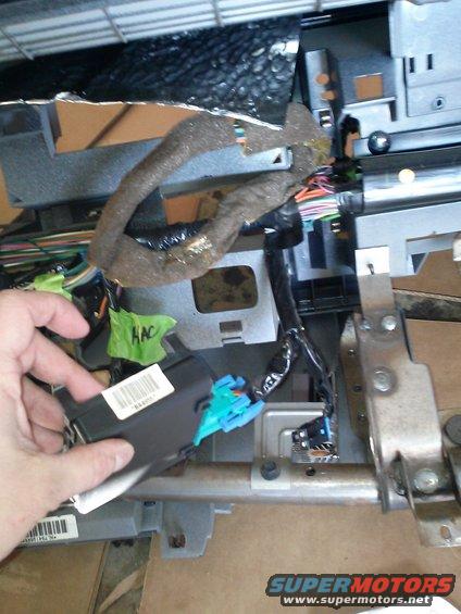Hand holding black control module with blue connector near vehicle dashboard wiring