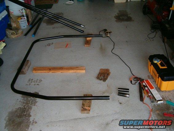 Black metal tubing, possibly for a roll cage, laid out on a garage floor with welding equipment and gloves