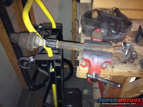 Driveshaft part clamped in a red vise, with yellow hose and black stand