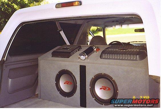 Interior shot of a vehicle showing a custom subwoofer box with two Alpine speakers, two amplifiers, and a capacitor
