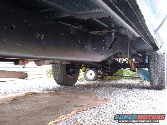Underside view of a truck's frame, suspension, and rear axle. A white wheelbarrow is visible in the background