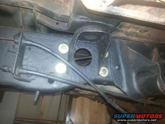 Underside of a vehicle frame with black painted suspension parts and brake lines. A circular opening is visible in a bracket