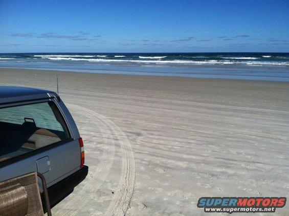 Silver station wagon parked on a sandy beach with ocean waves in the background. Tire tracks are visible in the sand