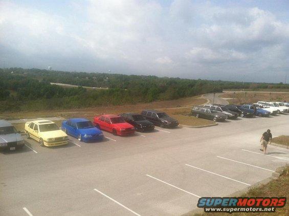 Several cars parked in a lot, including a yellow sedan, blue sedan, red sedan, and black SUVs