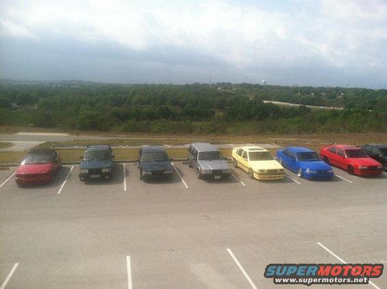 Several cars parked in a lot with a wooded area and cloudy sky in the background
