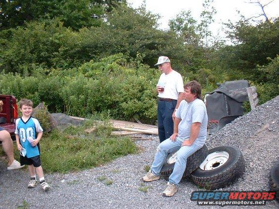 Kid in jersey, two men, and tires outside near trees and gravel