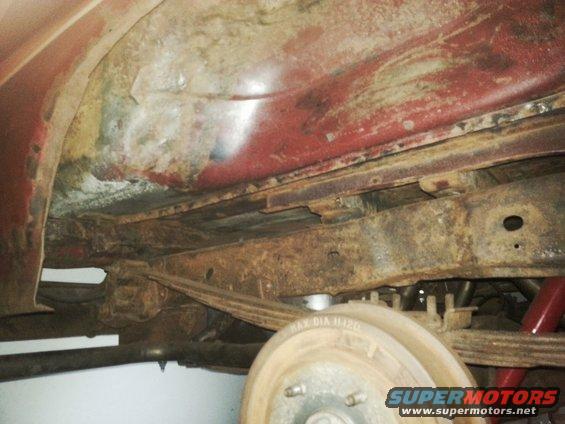 Underside of a red vehicle showing significant rust on the frame and body panels, with a rear drum brake and leaf spring visible