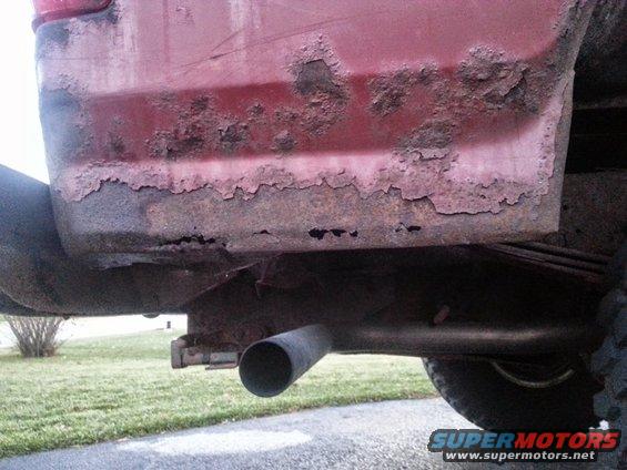 Red vehicle rear quarter panel with extensive rust damage and a visible exhaust pipe