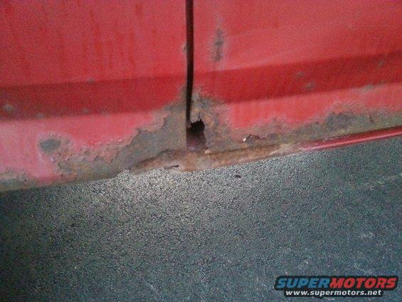 Red vehicle door seam with significant rust and a hole, close-up view