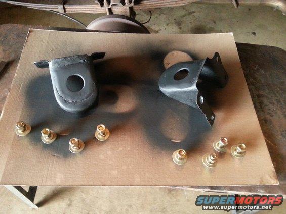 Two black metal brackets and gold nuts on cardboard, with Supermotors logo