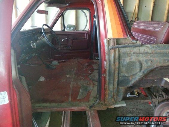 Interior of a red pickup truck, showing the driver's side door open, dashboard, steering wheel, and floor. The passenger side is disassembled
