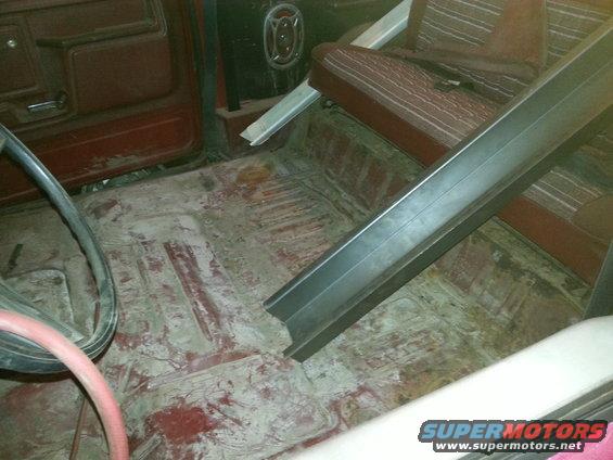 Interior shot of a vehicle with red seats and a bare metal floor, showing some rust and dirt