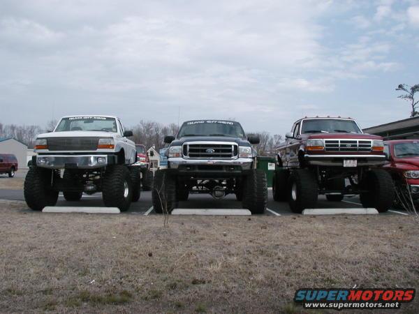 Three lifted pickup trucks parked side-by-side, front view. Red truck in foreground