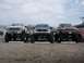 1985 Ford Bronco - Three lifted pickup trucks, including a red Ford F-Series, are parked in a lot under a cloudy sky.