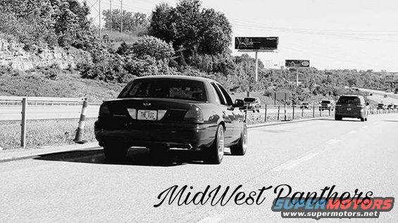Black 2004 Ford Crown Victoria driving on a highway, seen from behind. Other cars are on the road