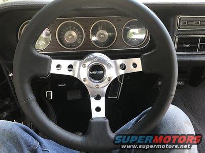 Black steering wheel with chrome spokes and a "NRB" logo, in front of a dashboard with three circular gauges